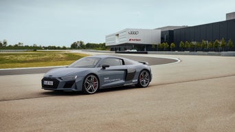 Matt gray Audi R8 Coupé with white Audi driving experience lettering on the driver's door driving on a track with an Audi Sport building in the background.