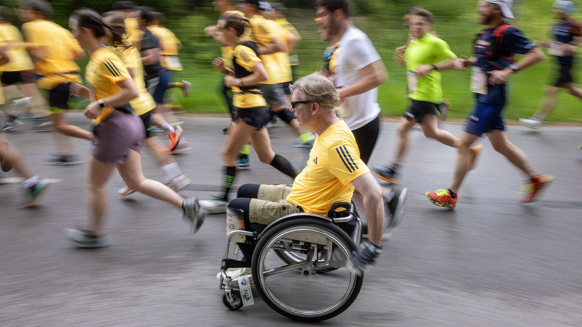 Mann im Rollstuhl mit schwarzer Sonnenbrille im gelben Laufshirt auf nasser Straße, umgeben von laufenden Teilnehmenden in Gelb.
