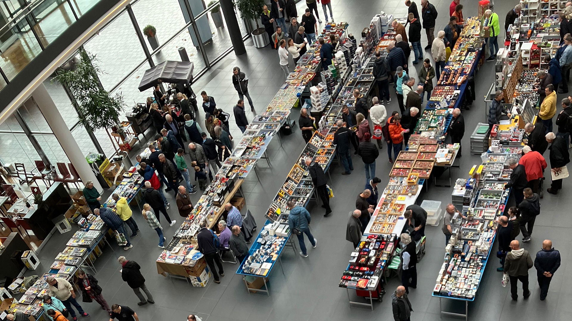 Draufsicht auf die Modellautoboerse 2023 im Audi Forum Neckarsulm. Aufgereihte Tische im Foyer des Audi Forums präsentieren Modellautos zwischen den Tischen gehen Menschen und schauen sich die Modelle an.