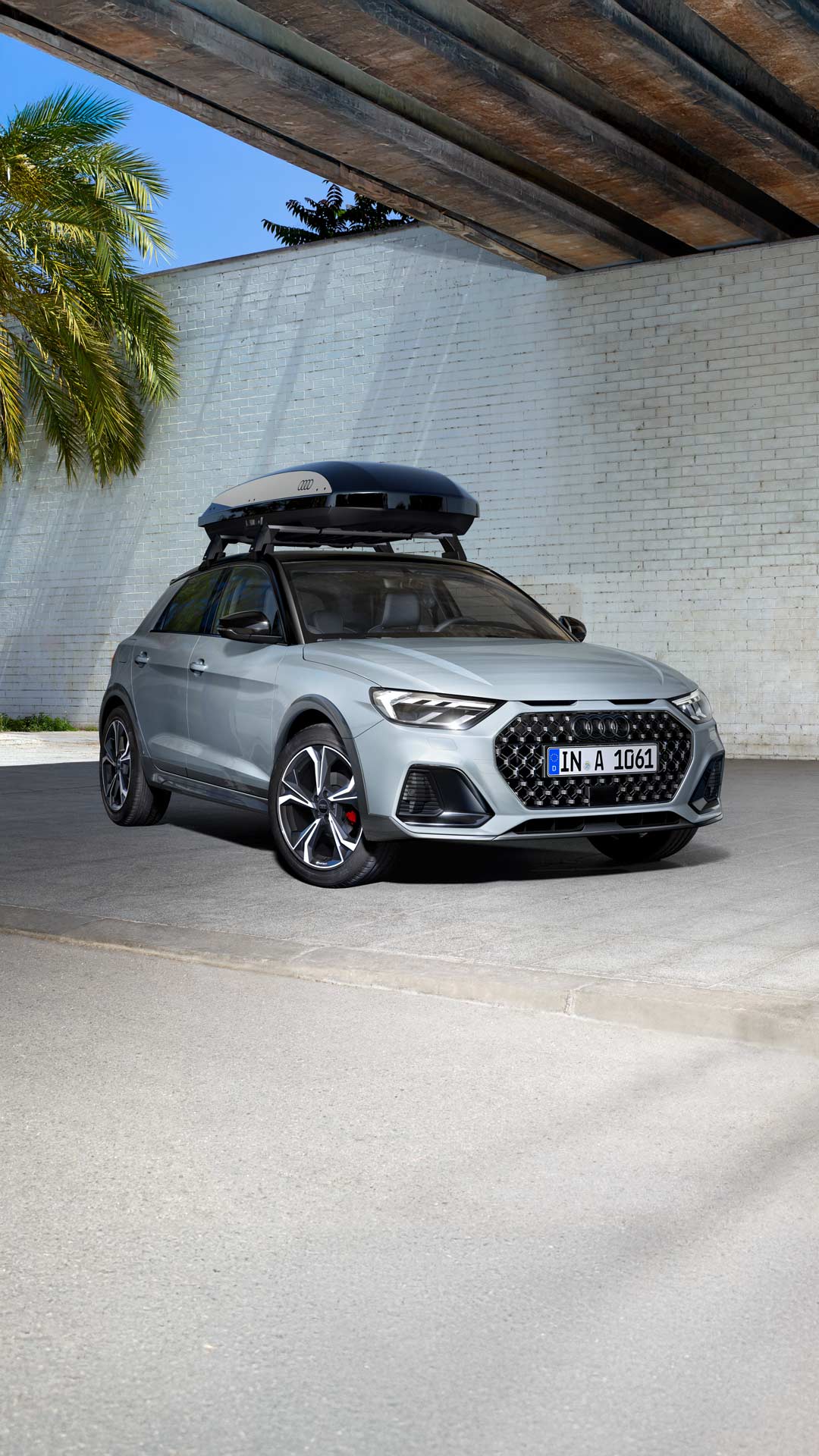 Silberner Audi A1 allstreet mit Dachbox vor weißer Ziegelwand auf grauem Pflaster geparkt.