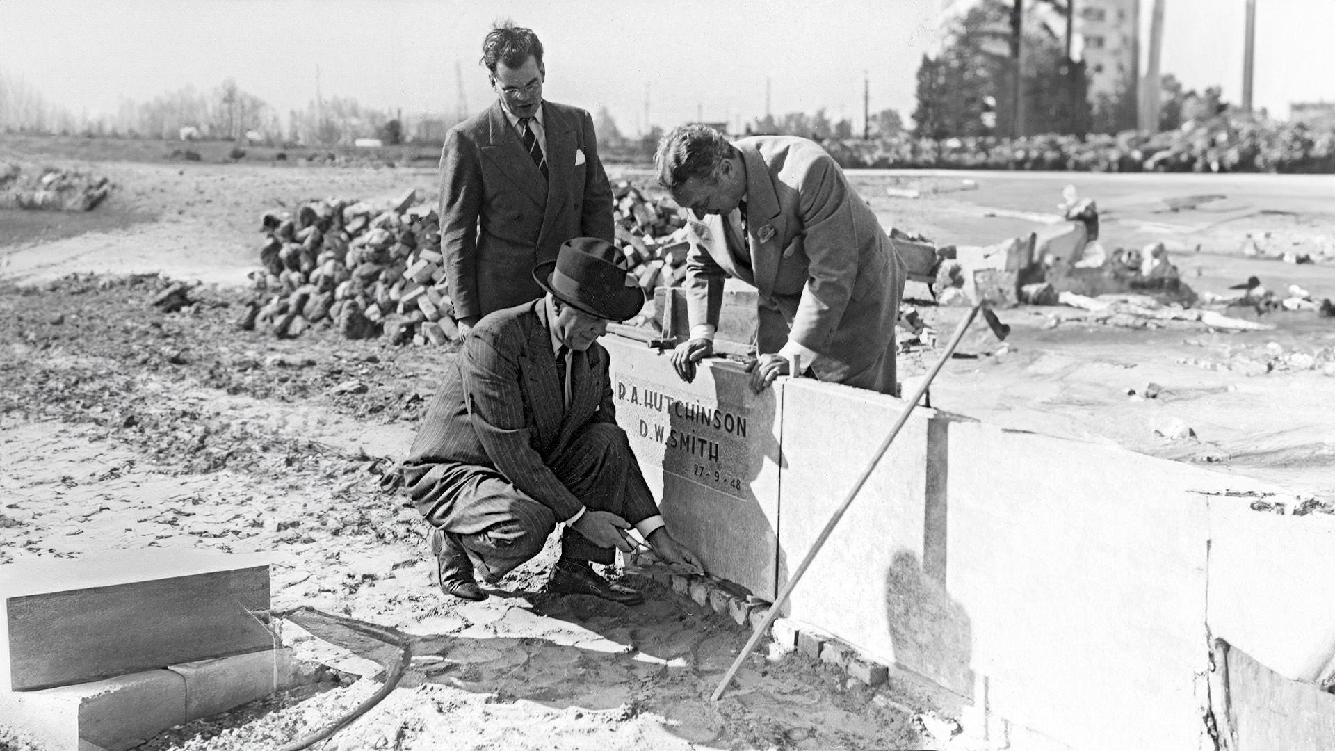 Une vieille photo en noir et blanc montrant 3 hommes inspectant un bâtiment sur le côté