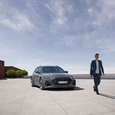 Mattgrauer Audi RS 6 Avant performance auf heller Fläche, Mann im Anzug geht rechts am Fahrzeug vorbei.