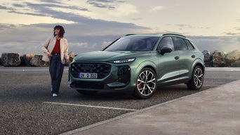 Der Q3 SUV schräg von vorn auf einem Parkplatz, im Hintergrund begrenzt von Wackersteinen. Eine modisch gekleidete junge Frau bewegt sich neben dem Fahrzeug.