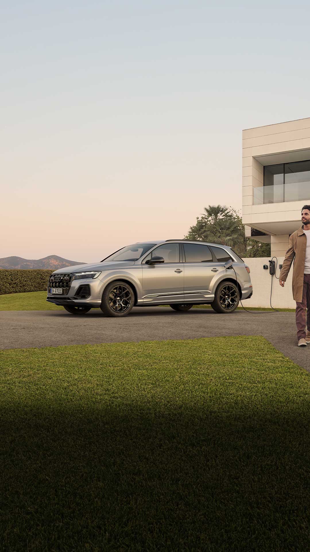 Ein silberner Audi Q7 SUV TFSI e lädt an Wallbox vor modernem Haus, ein lächelndes Paar im Businessoutfit läuft Arm in Arm rechts am Auto vorbei.
