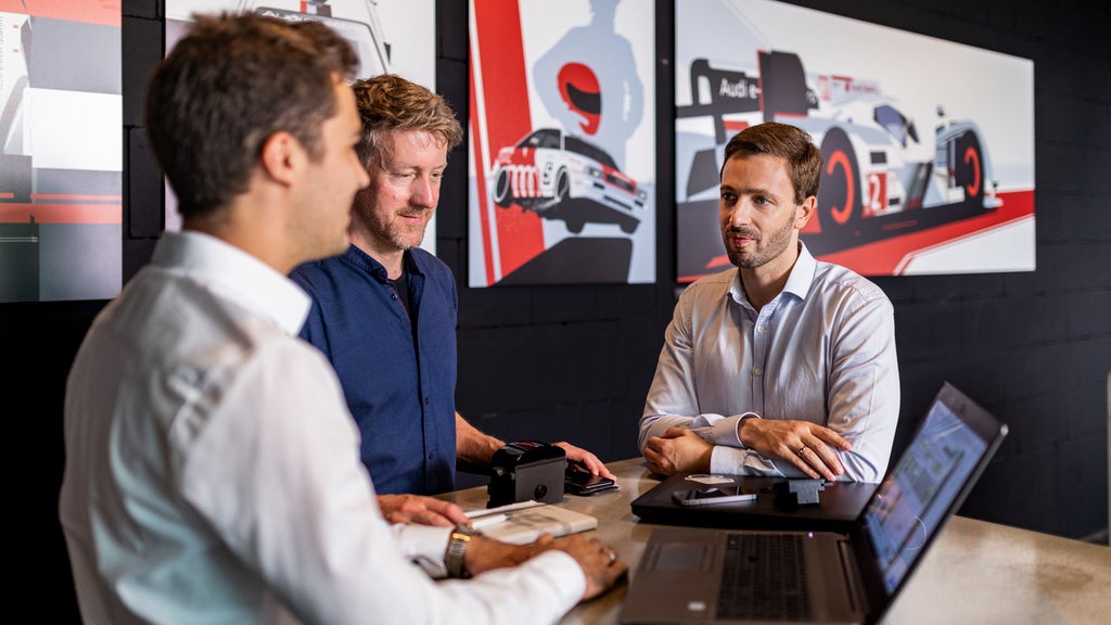 Three men are sitting around a table where a laptop is opened