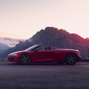 Roter Audi R8 Spyder V10 mit offenem Verdeck steht vor imposanter Bergkulisse bei Sonnenuntergang auf einer asphaltierten Straße.