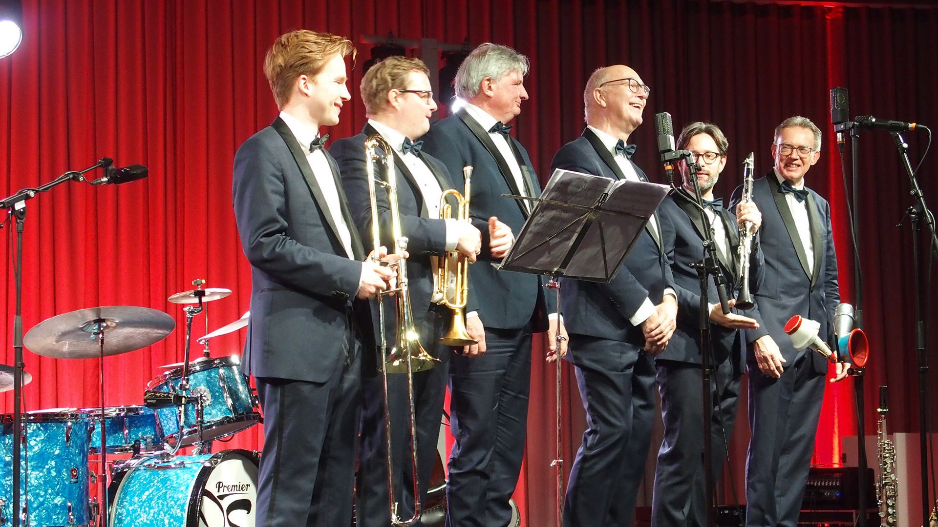 Mehere Männer in Anzug mit Musikinstrumenten stehen vor einem roten Vorhang auf einer Bühne, ein Mann singt, im Hintergrund sieht man ein Schlagzeug mit Trommeln.