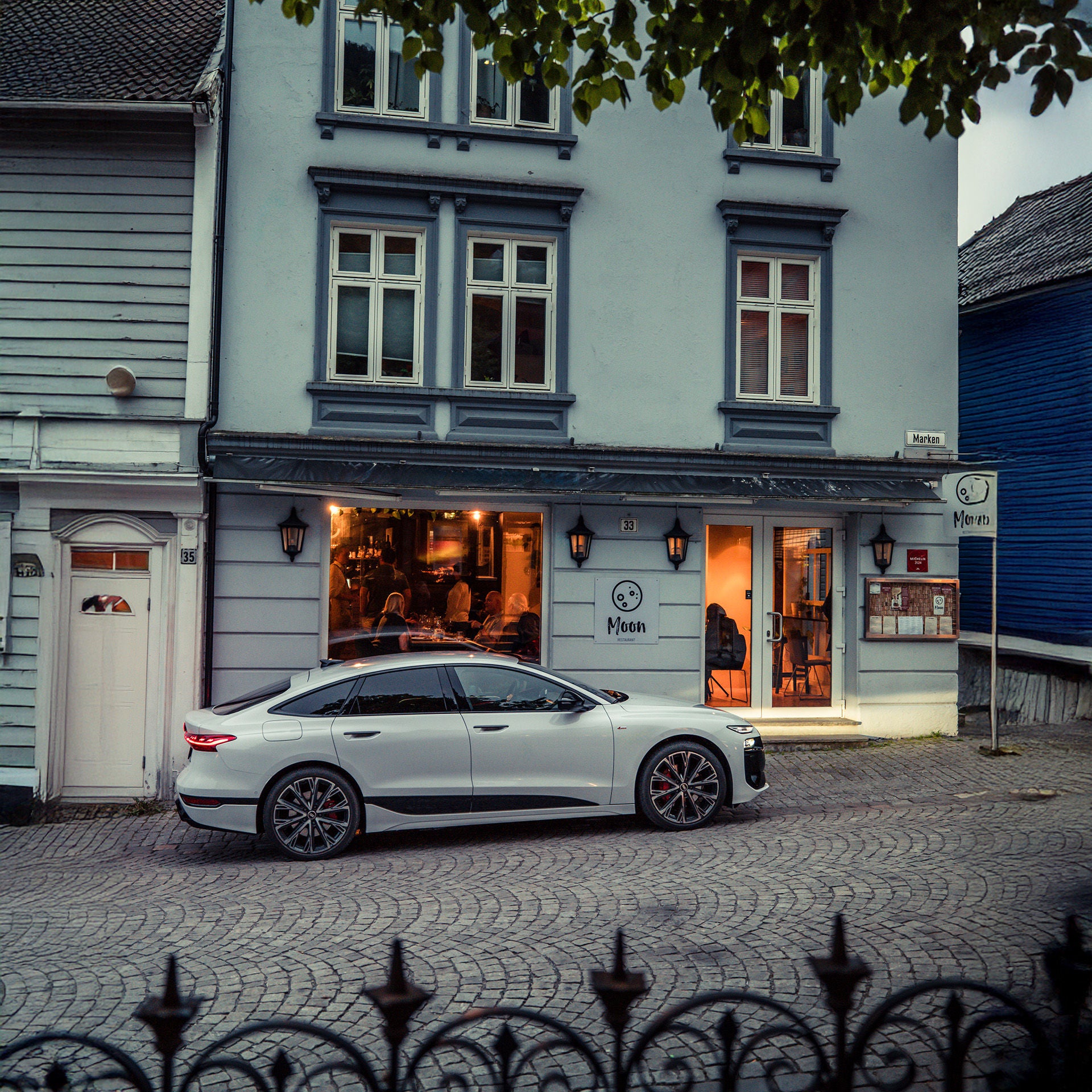 The white Audi A6 Sportback e-tron performance stands at the end of the journey in front of a house in Bergen. The profile view emphasises the focus on dynamism, elegance and progressiveness of its design.