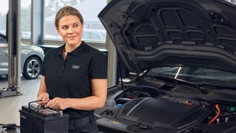 Frau mit Batterie in der Hand steht vor einem Auto, wo die Motorhaube offen ist