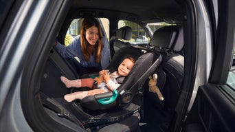 Lächelnde Frau schaut auf ein Baby im Kindersitz auf dem Rücksitz eines Audi, das ein Kuscheltier hält.
