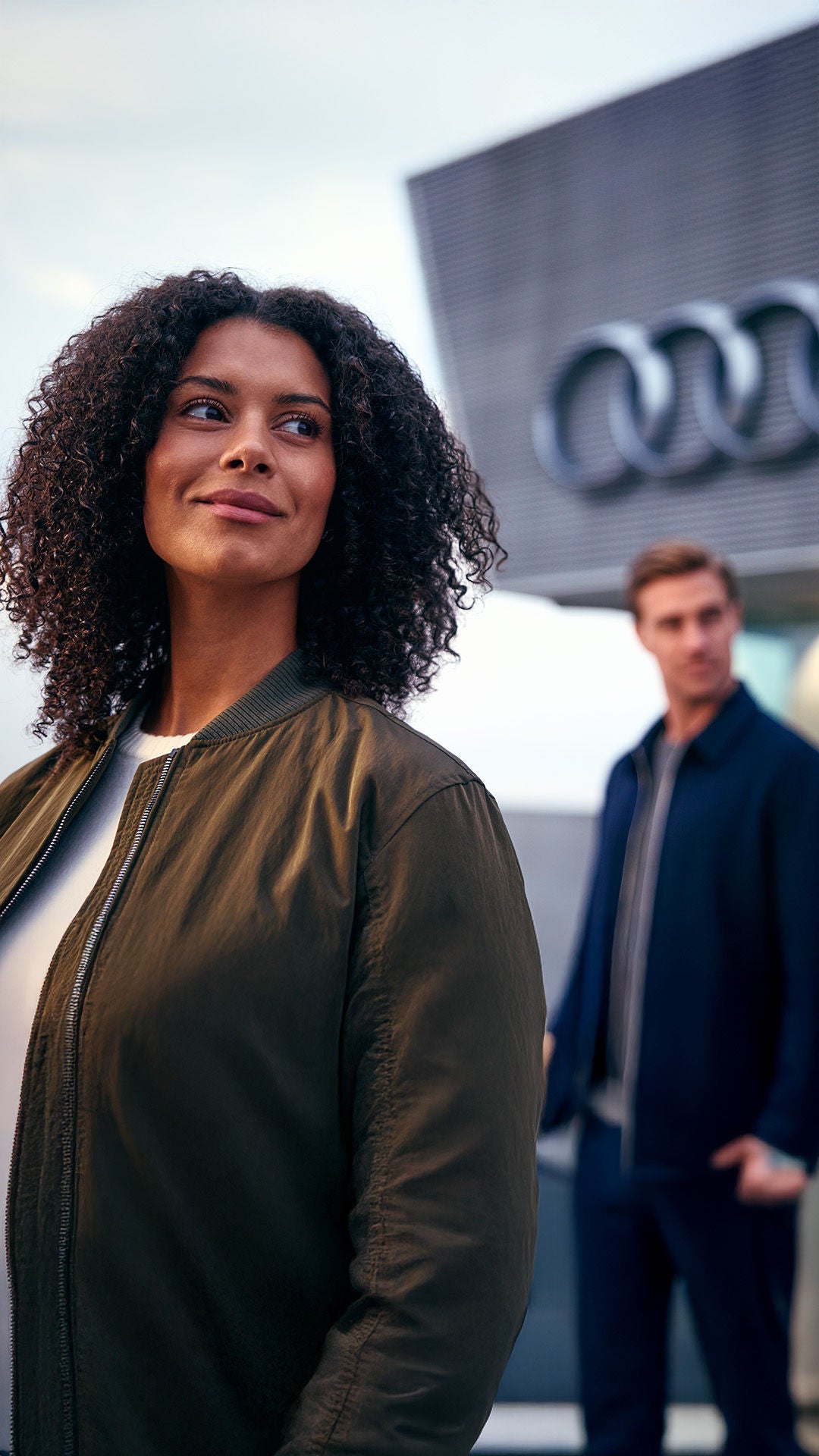Eine Frau mit lockigem Haar steht vor einem Gebäude mit Audi-Logo, ein Mann steht im Hintergrund und blickt zu ihr.