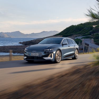 Dunkelblauer Audi S6 Avant e-tron fährt an einer Küstenstraße entlang, im Hintergrund Meer und Hügel.