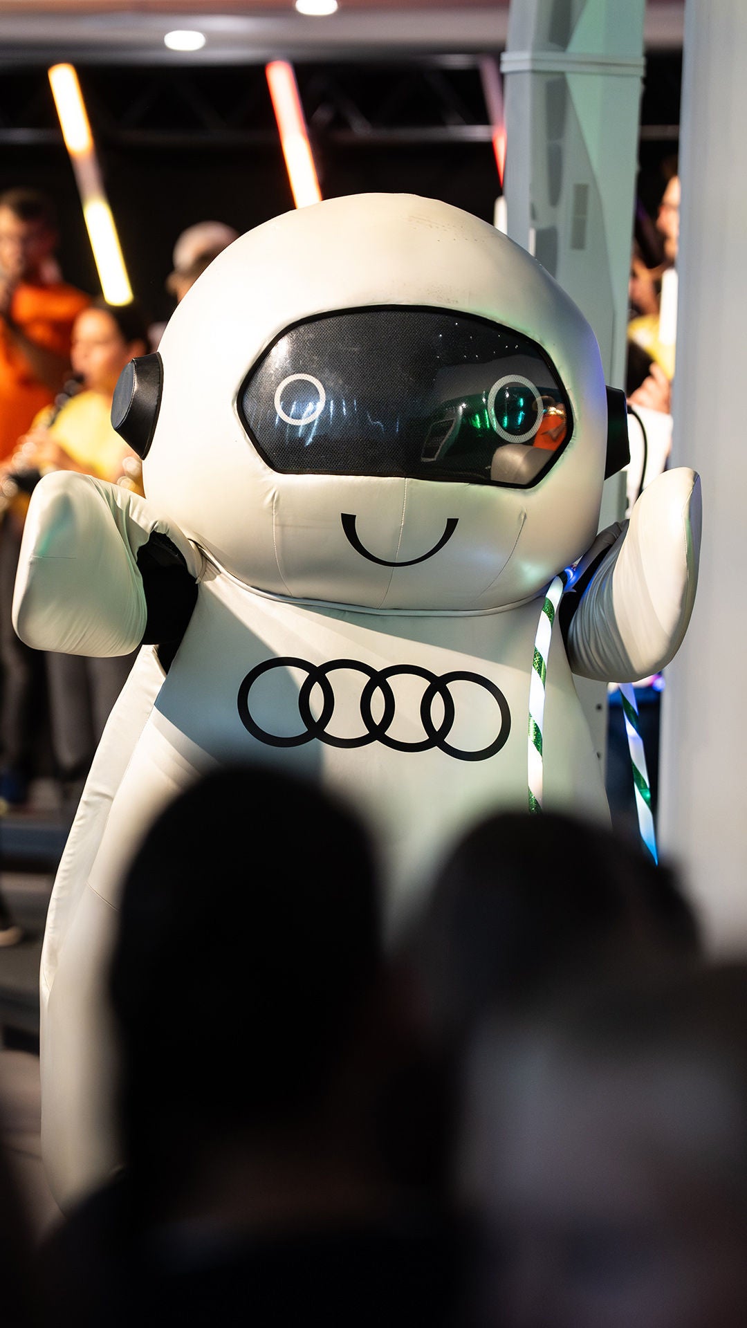 Weißes Audi Robotermaskottchen ADUI vor einem Orchester mit bunten Leuchtstäben bei einem Kinderkonzert.