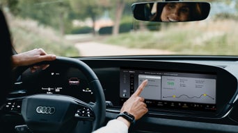 Fahrerin bedient großes Autodisplay mit Fahrdaten; eine Hand am Lenkrad, ihr Gesicht im Rückspiegel sichtbar