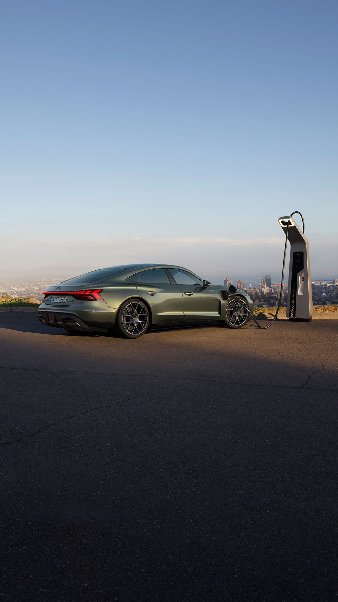 A green electric car charging at a modern station, overlooking an urban skyline under a clear sky.