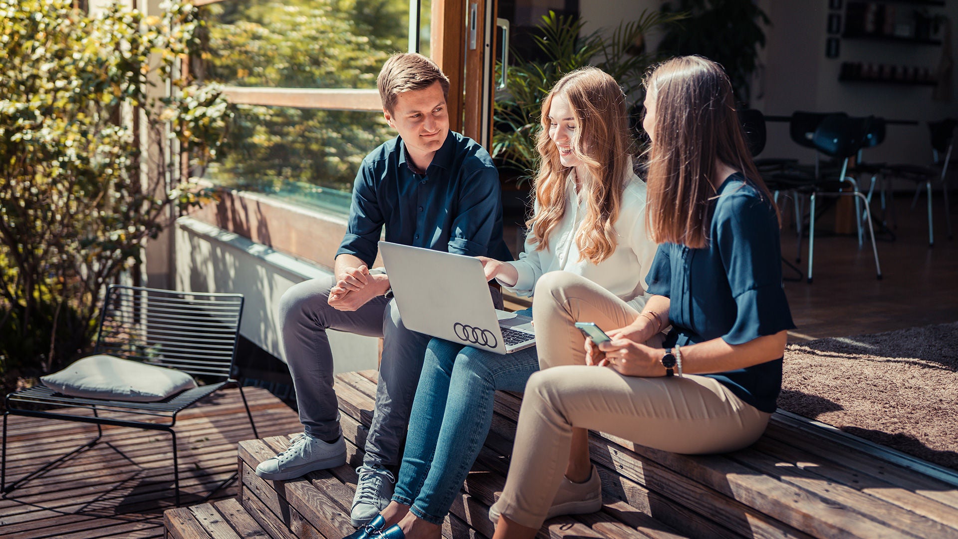 Three people are sitting together chatting in the sunshine with laptop on lap. Basic Teaser