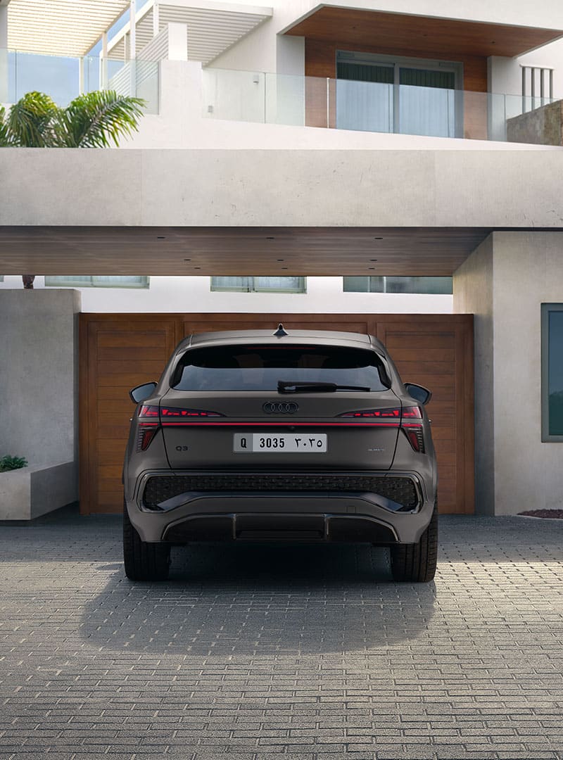 The Audi Q3 Sportback can be seen from the rear view. It is parked in front of a wooden entrance gate to a residential building.