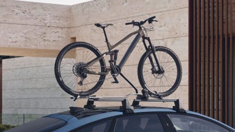 Mountainbike ist auf Dachträger eines blauen Audi montiert, im Hintergrund moderne Stein- und Holzarchitektur.