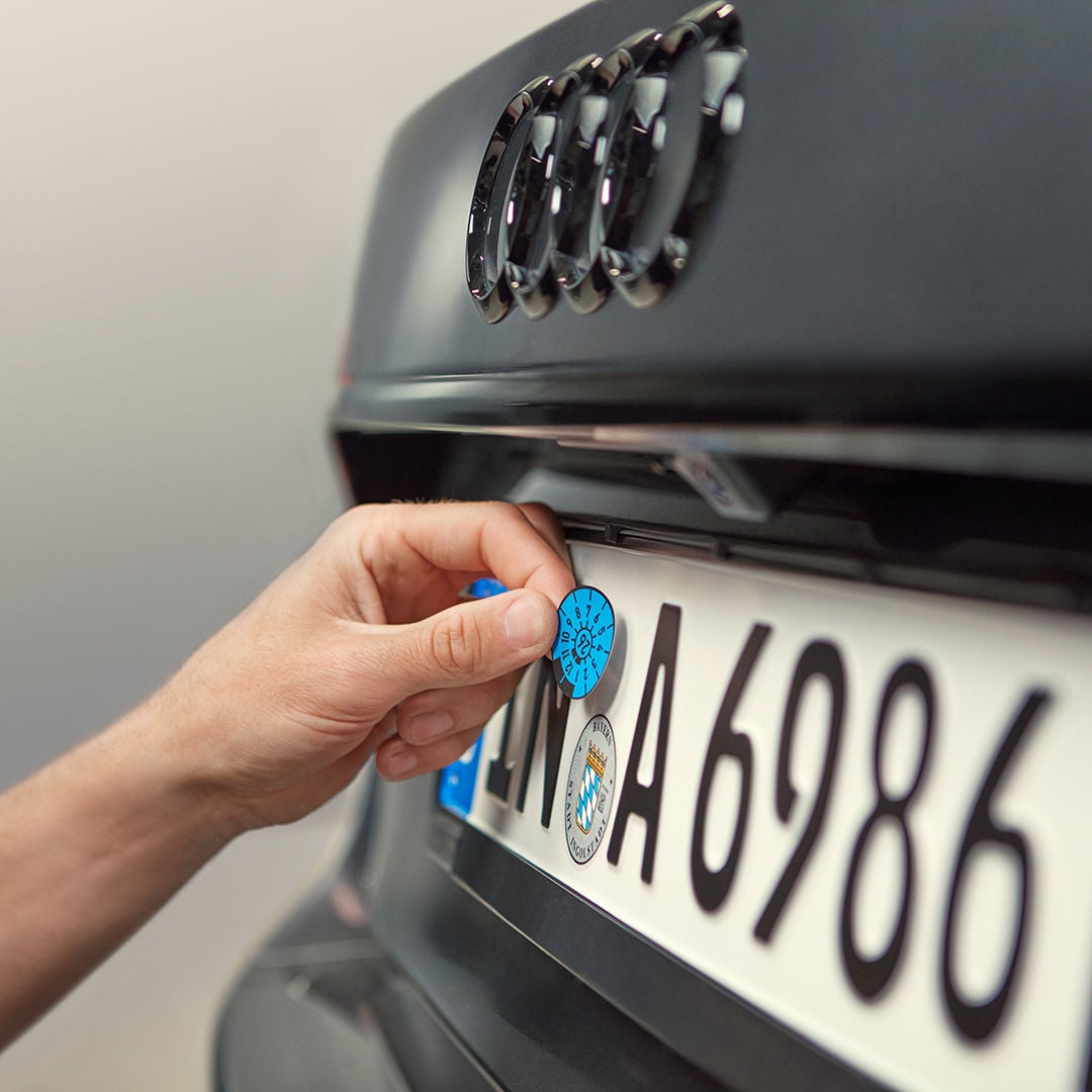 Hand bringt blaue Prüfplakette an einem deutschen Autokennzeichen unter dem Audi-Logo an, Fokus auf Nahaufnahme.