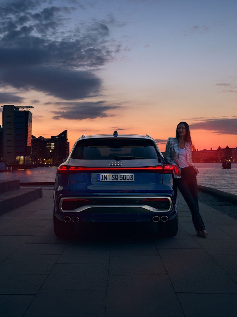 Ein blauer Audi SQ5 SUV bei Sonnenuntergang am Wasser, Frau steht daneben vor moderner Stadtkulisse.