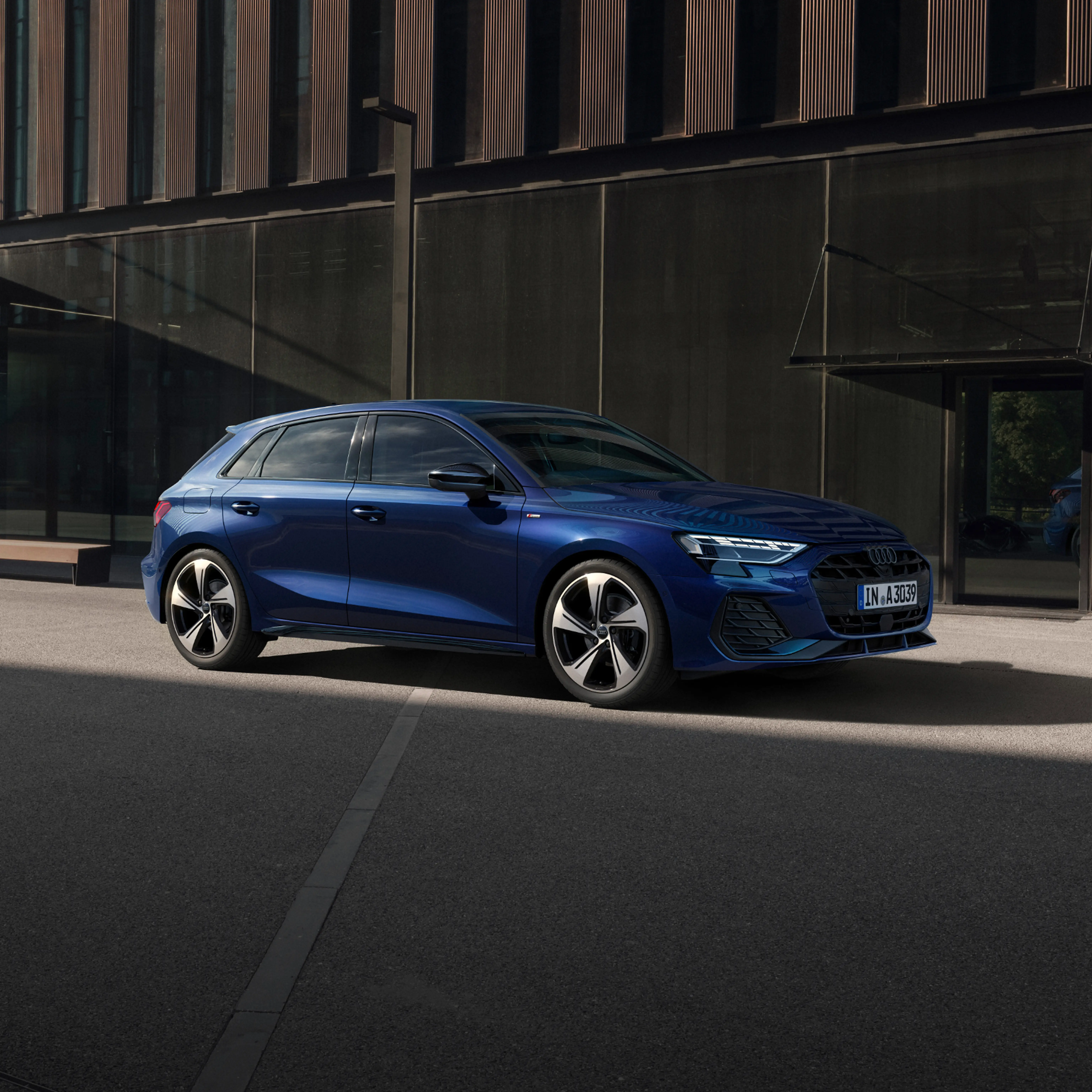 A green Audi A3 is parked on a sunlit city street, casting a shadow on the pavement. The modern building in the background adds a stylish, urban feel.