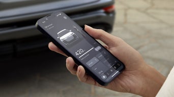 Die Hand einer Frau ragt von rechts ins Bild. Sie hält ein Smartphone in der Hand, auf dessen Display Funktionen zum Q6 e-tron angeboten werden. Im Hintergrund steht unscharf ein grüner Audi Q6 e-tron.