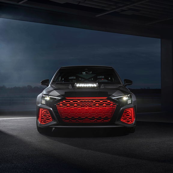 Schwarz/grauer Audi RS 3 driving experience mit orangefarbener Front und Zusatzscheinwerfer steht frontal in dunkler Halle.  