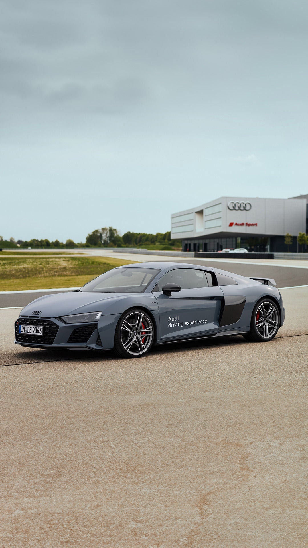 A matte gray Audi R8 Coupé V10 performance quattro drives along a track, with an Audi Sport building in the background.