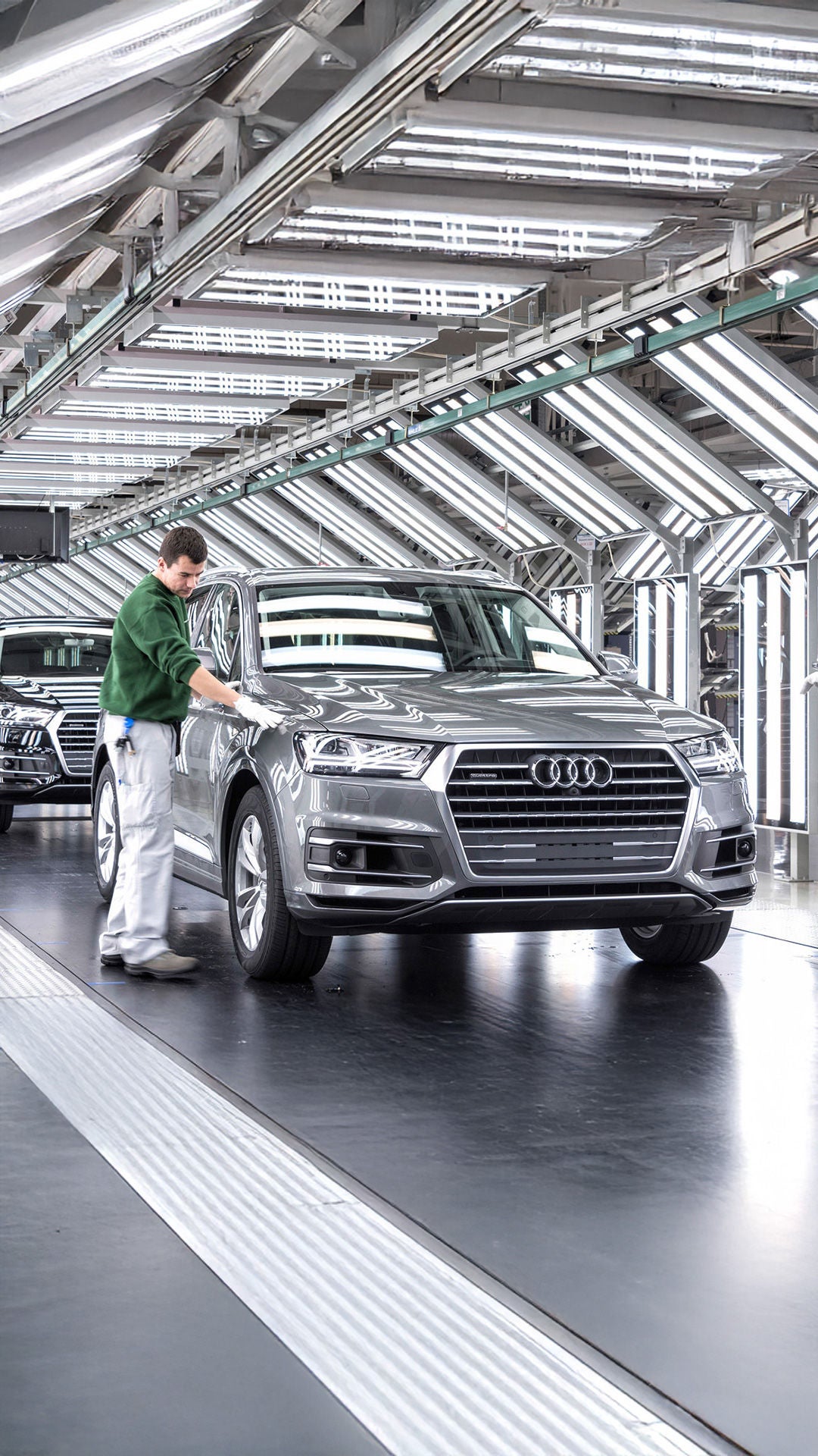 Zwei Mitarbeitende inspizieren einen grauen Audi Q7 in einer modernen Produktionslinie mit LED-Beleuchtung.