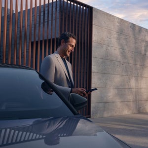 A man in a grey blazer smiles while using a smartphone beside a sleek car. The modern setting features a concrete wall and wooden accents, creating a serene, sophisticated atmosphere.