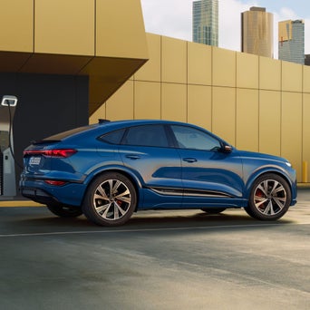The Q4 Sportback e-tron is parked in front of a modern building façade and is connected to a charging station.