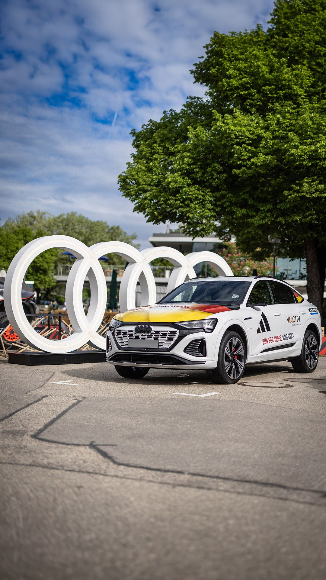 Weißer Audi Q8 e-tron 55 quattro mit Deutschlanddesign vor Audi Ringen; links ein ehemaliges Rallye-Dakar-Rennfahrzeug.
