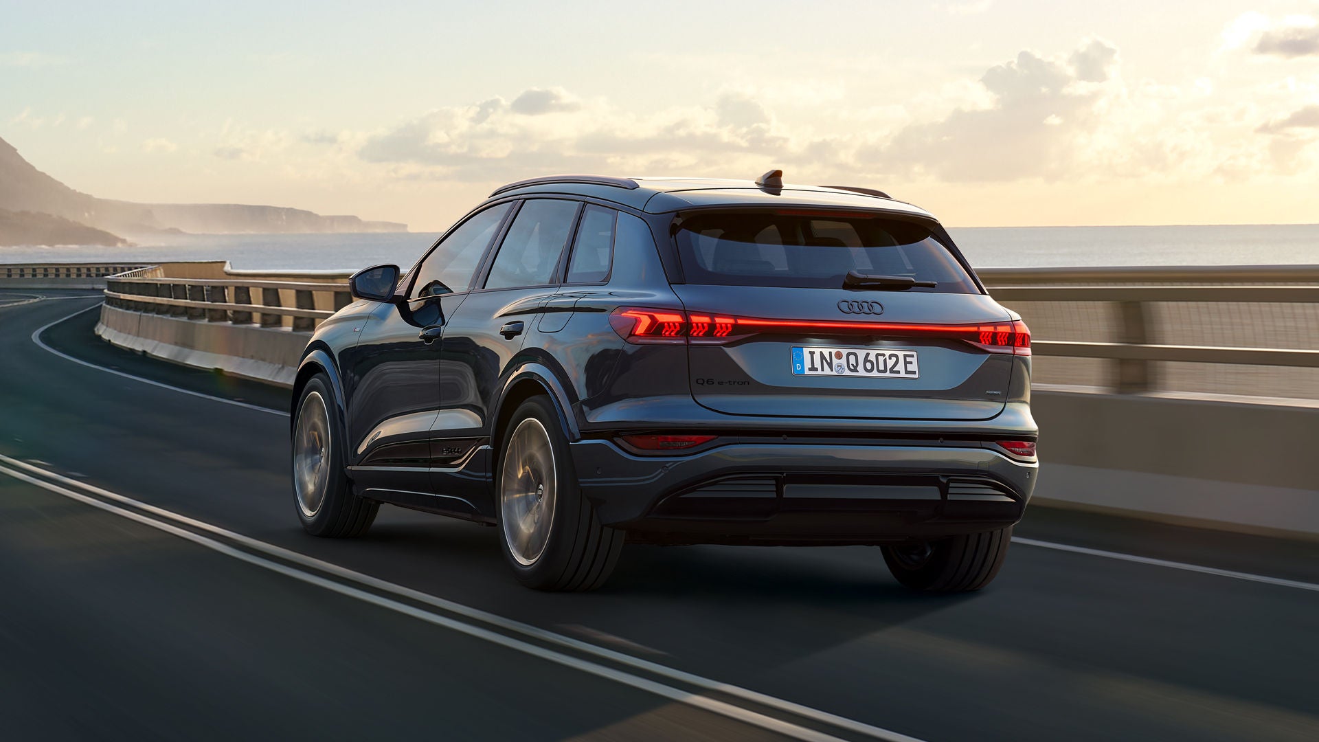 Schräge Heckaufnahme des Q6 SUV e-tron, welche über eine kurvige Straße am Meer fährt. Im Hintergrund sind Berge zu sehen.