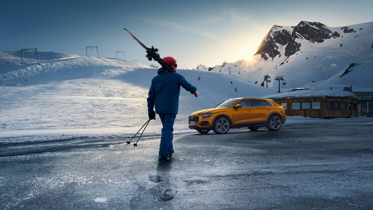 Someone skis towards an orange Audi in a mountain landscape.