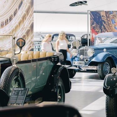 Two women look at vintage cars in a bright museum exhibition of historic vehicles.