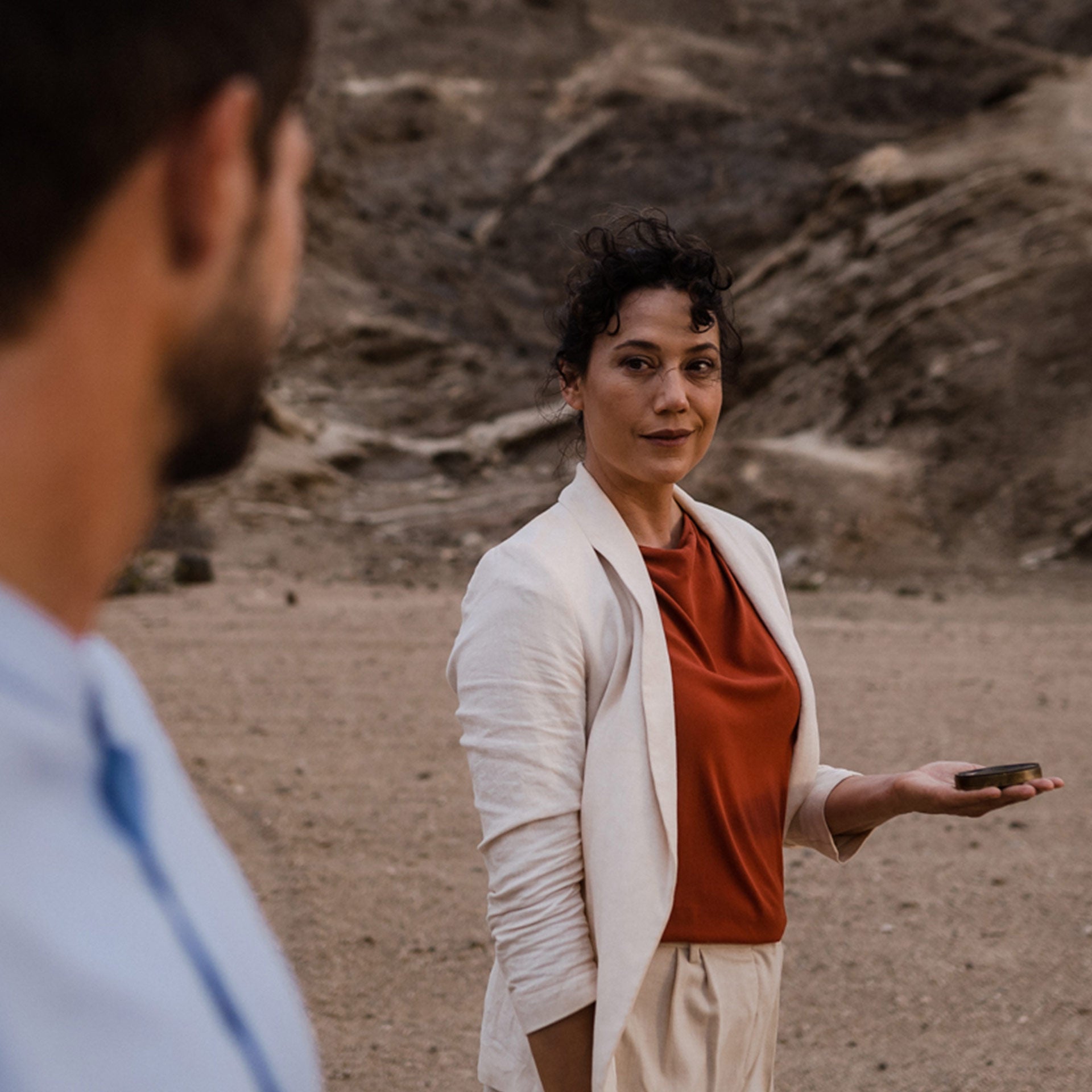 A woman stands in a desert landscape and looks into the eyes of a man in the foreground. A compass lies on her outstretched left hand.