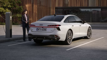 Die A6 Limousine e-hybrid steht dem Eingangsbereich eines großen Gebäudes zugewandt. Links vom Heck des Fahrzeugs steht eine Frau mit einem Ladekabel in der Hand und blickt auf das Fahrzeug.
