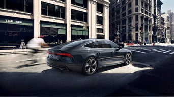 Ein grauer Audi A7 fährt durch eine Straßenecke in der Stadt, Fahrradfahrer unscharf im Vordergrund, Gebäude im Hintergrund.