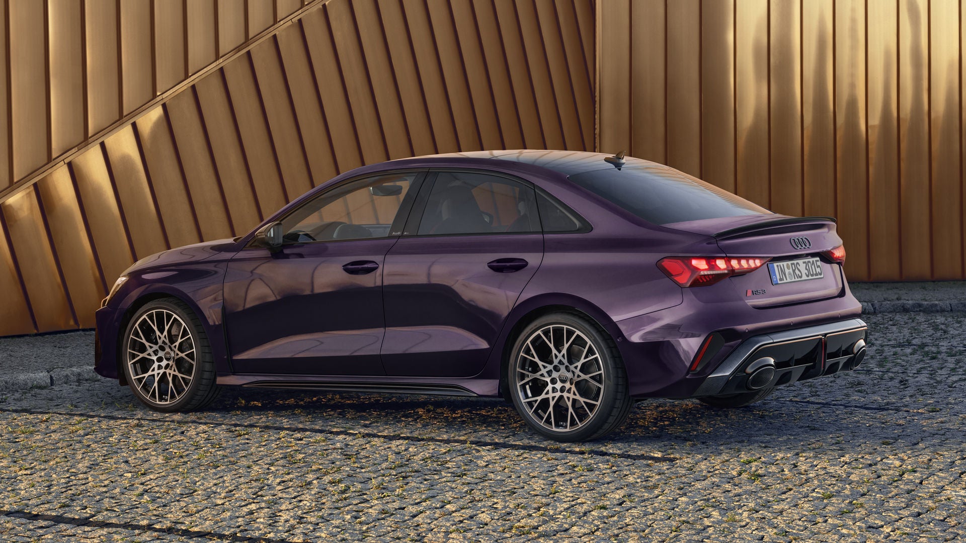 Die RS 3 Limousine mit Indivuduallackierung in Merlin Perleffekt von der Seite parkend auf Kopfsteinpflaster, im Hintergrund Umrisse eines modernen Gebäudes.