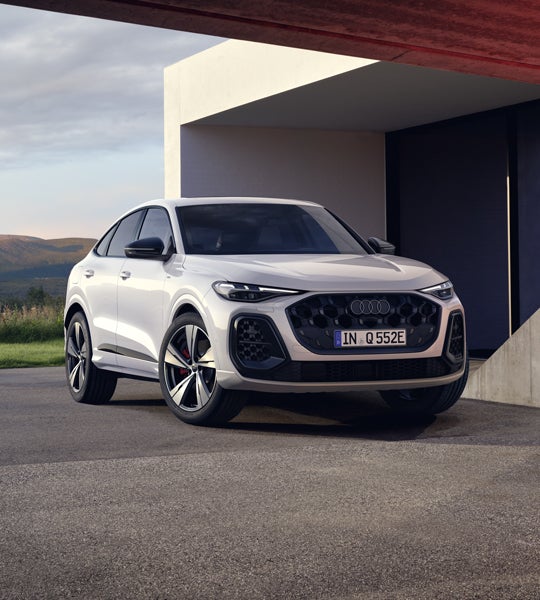Der weiße Q5 Sportback e-hybrid in schräger Frontansicht steht vor einem modernen Haus. Im Hintergrund sieht man eine bergige Landschaft.