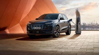 Ein grauer Audi SQ6 SUV e-tron wird an einer Ladesäule vor moderner Architektur mit Stadtblick bei Sonnenuntergang geladen.