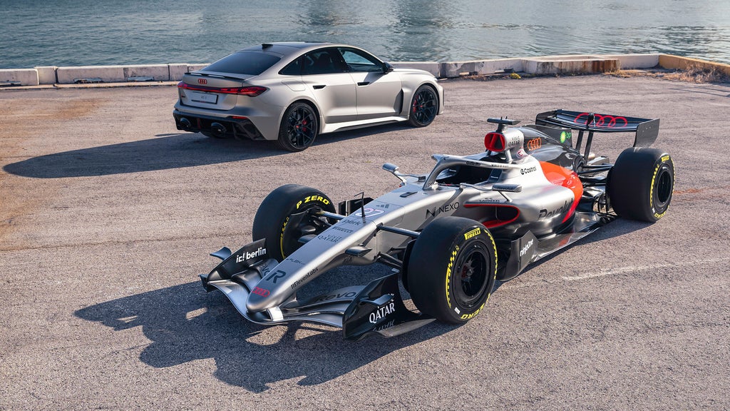 Silberner Formel-1-Wagen Audi R26 mit roten Akzenten steht auf Asphalt am Wasser; dahinter ein grauer Audi RS 5 Avant.