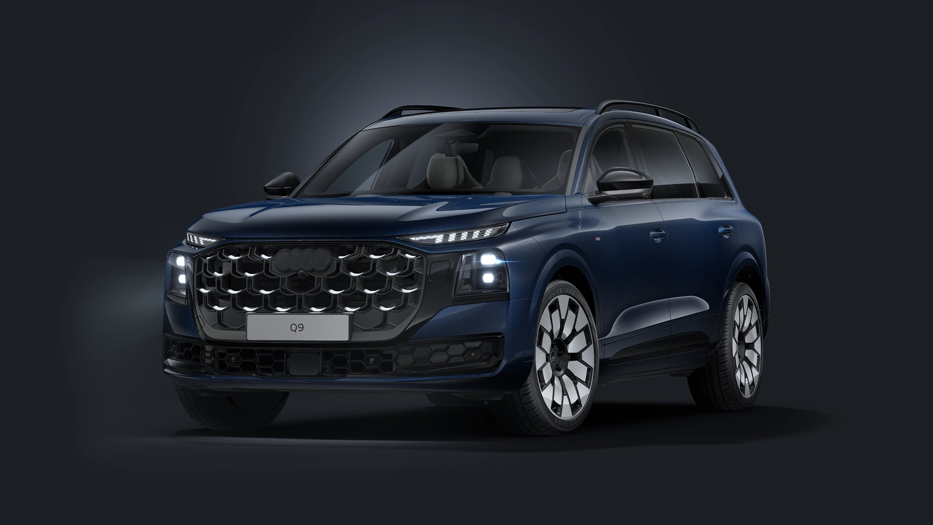 Blue Audi Q9 SUV shown in a studio setting from a front three-quarter angle, featuring a prominent grille, illuminated headlights, and alloy wheels against a dark background.
