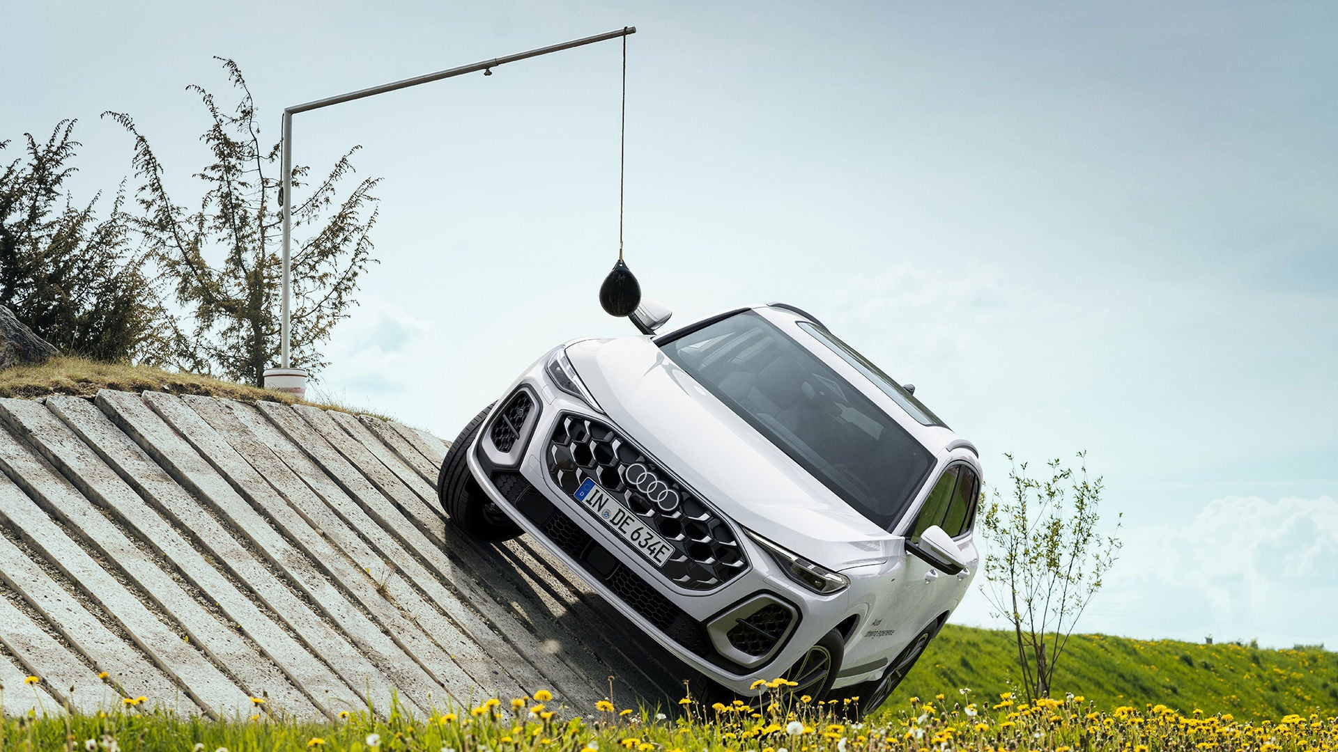 Weißer Audi Q5 SUV e-hybrid auf schräger Betonrampe; Frontansicht mit Audi-Logo, Blumenwiese und Himmel im Hintergrund.