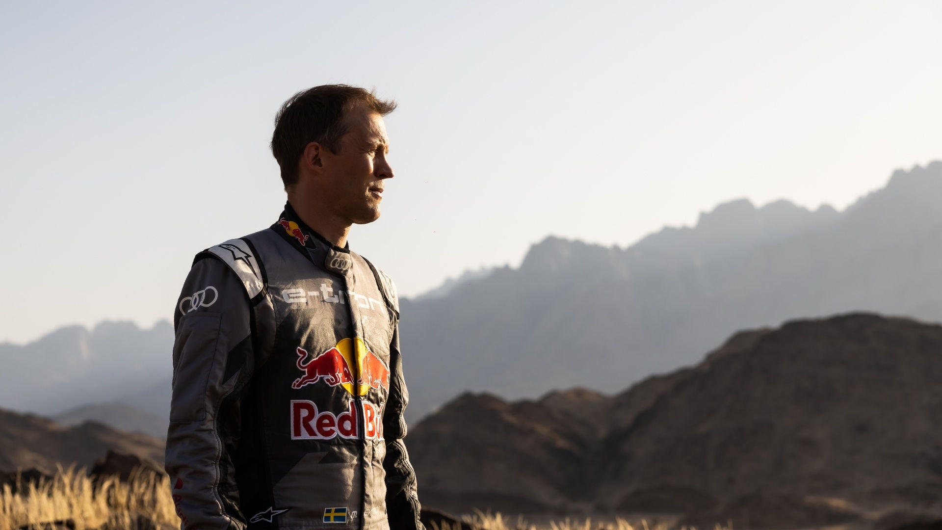 Rallyefahrer im Red-Bull-Anzug steht vor felsiger Berglandschaft, blickt in die Ferne bei Sonnenlicht.