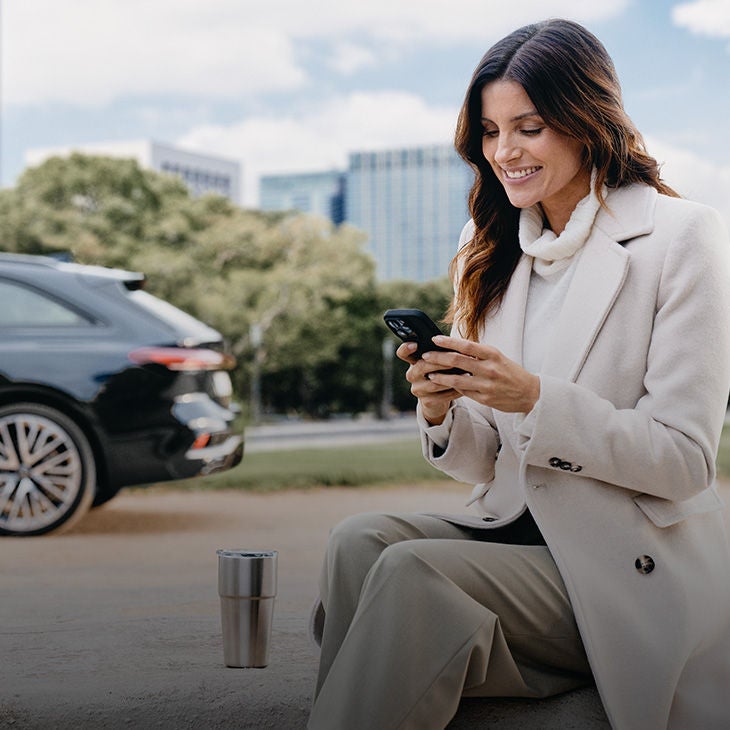 Lächelnde Frau sitzt im Freien, trägt einen weißen Mantel und benutzt ein Smartphone. Im Hintergrund steht ein schwarzer Audi vor einer urbanen Kulisse mit Bäumen und Bürogebäuden.