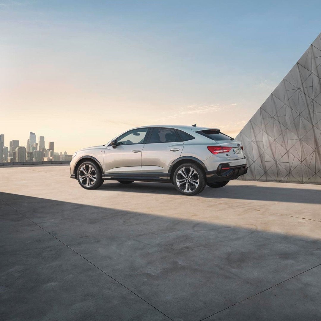 Silver Audi Q3 Sportback parked on a rooftop with city skyline and geometric metallic wall at sunset.