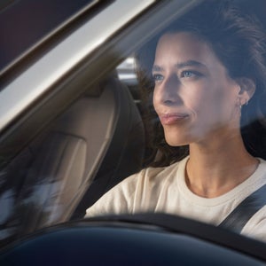 Woman with curly hair driving a car, viewed through the windshield. She wears a seatbelt and a light sweater, looking forward calmly and confidently.