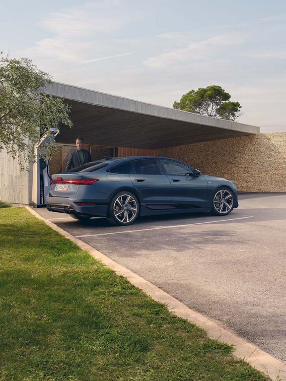 Blauer Audi A6 Sportback e-tron lädt an Wallbox vor moderner Hausfassade, ein Mann steht daneben.