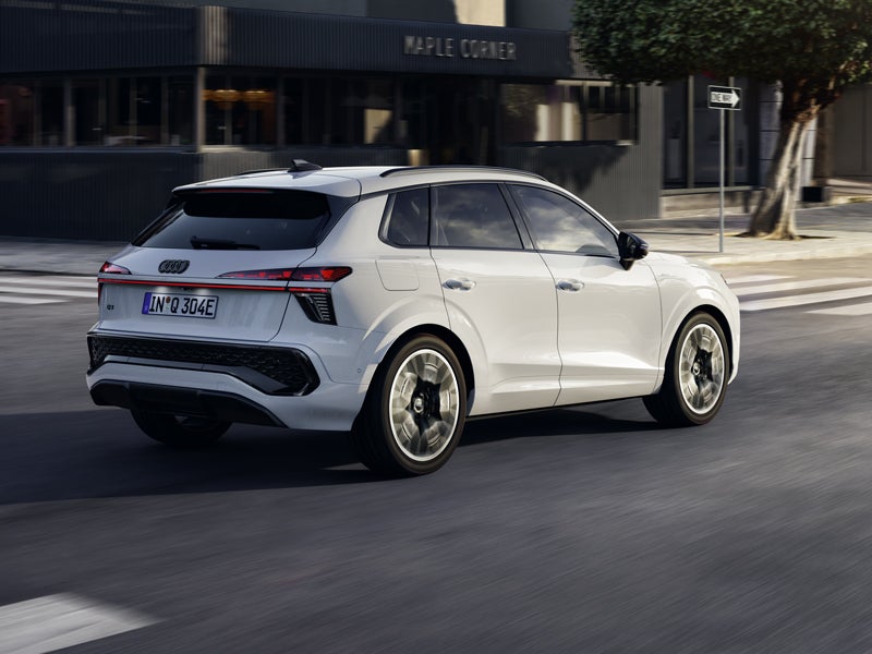 Ein weisser Q3 SUV e-hybrid, von hinten rechts gesehen, während er an einer Kreuzung in der Innenstadt fährt.